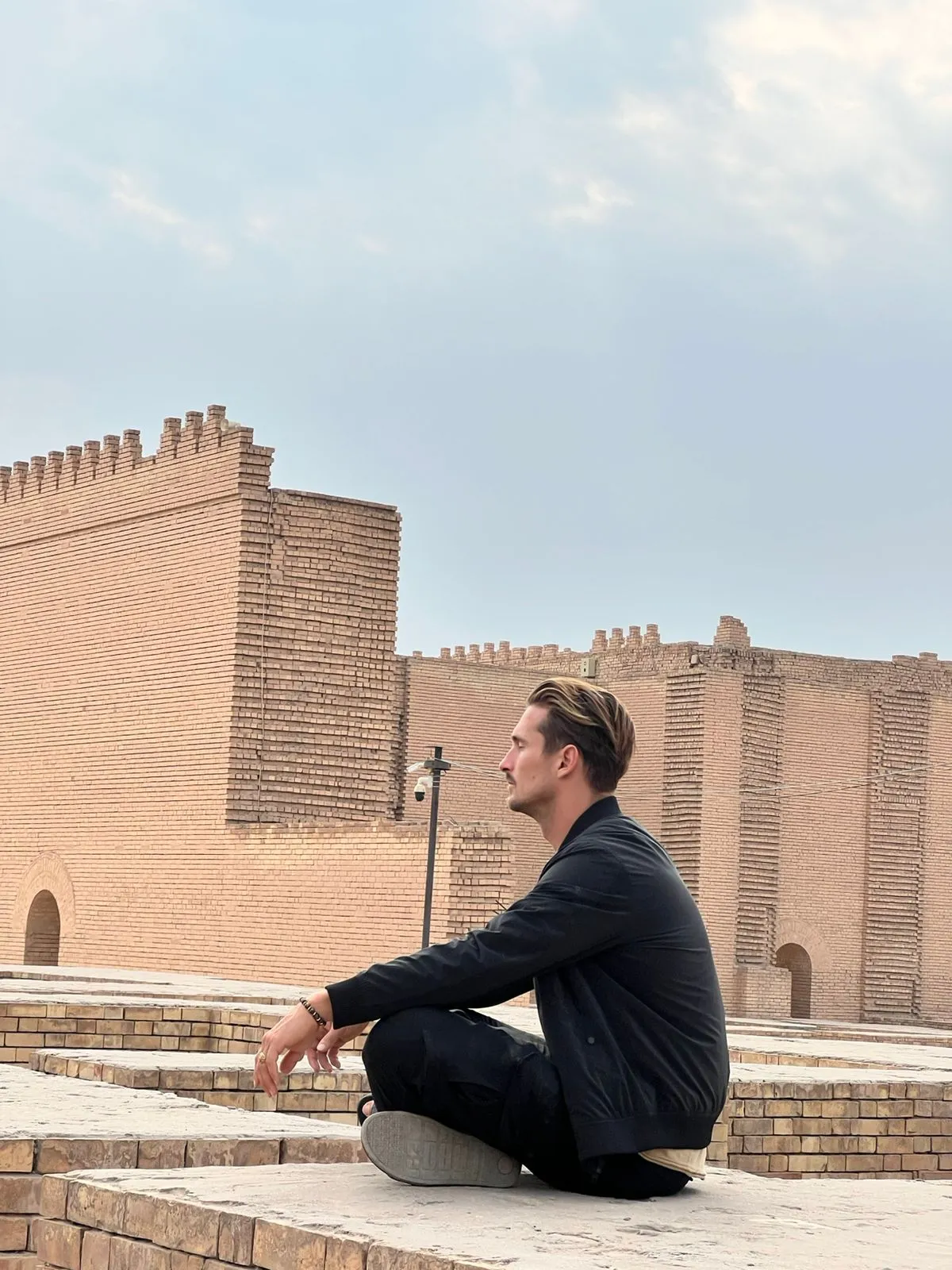 A visitor sitting on the brick steps at Babylon, the reconstructed walls and crenellated towers of the city rising behind him.