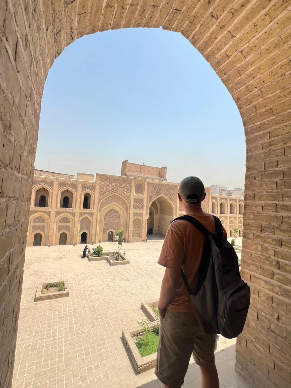 The courtyard of the Al-Mustansiriya Madrasa in Baghdad framed by a brick archway, its carved iwan and tiers of arched cells facing across the open paving.