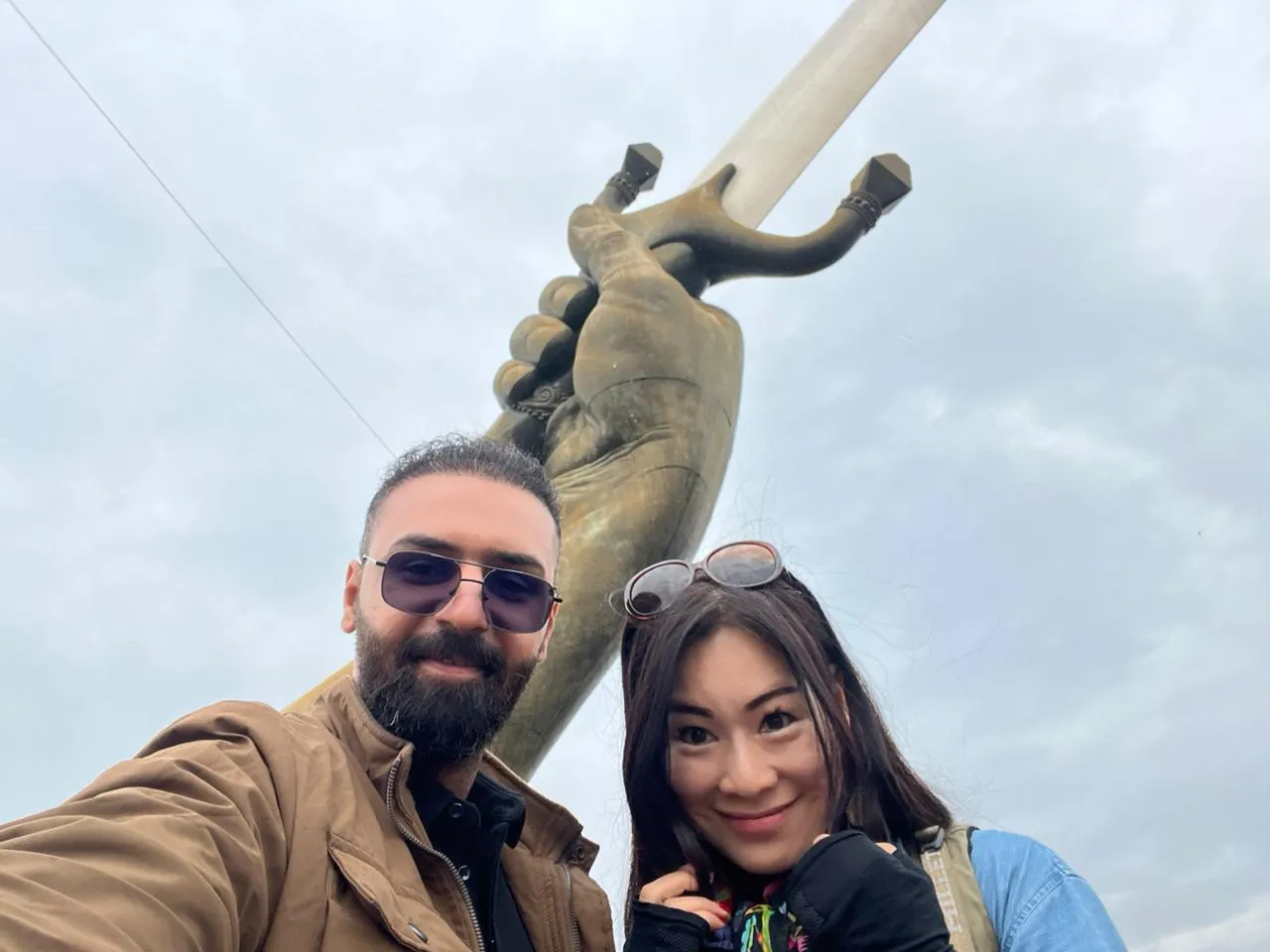 A bronze monument photographed from below against a pale sky, two travellers smiling in the foreground.