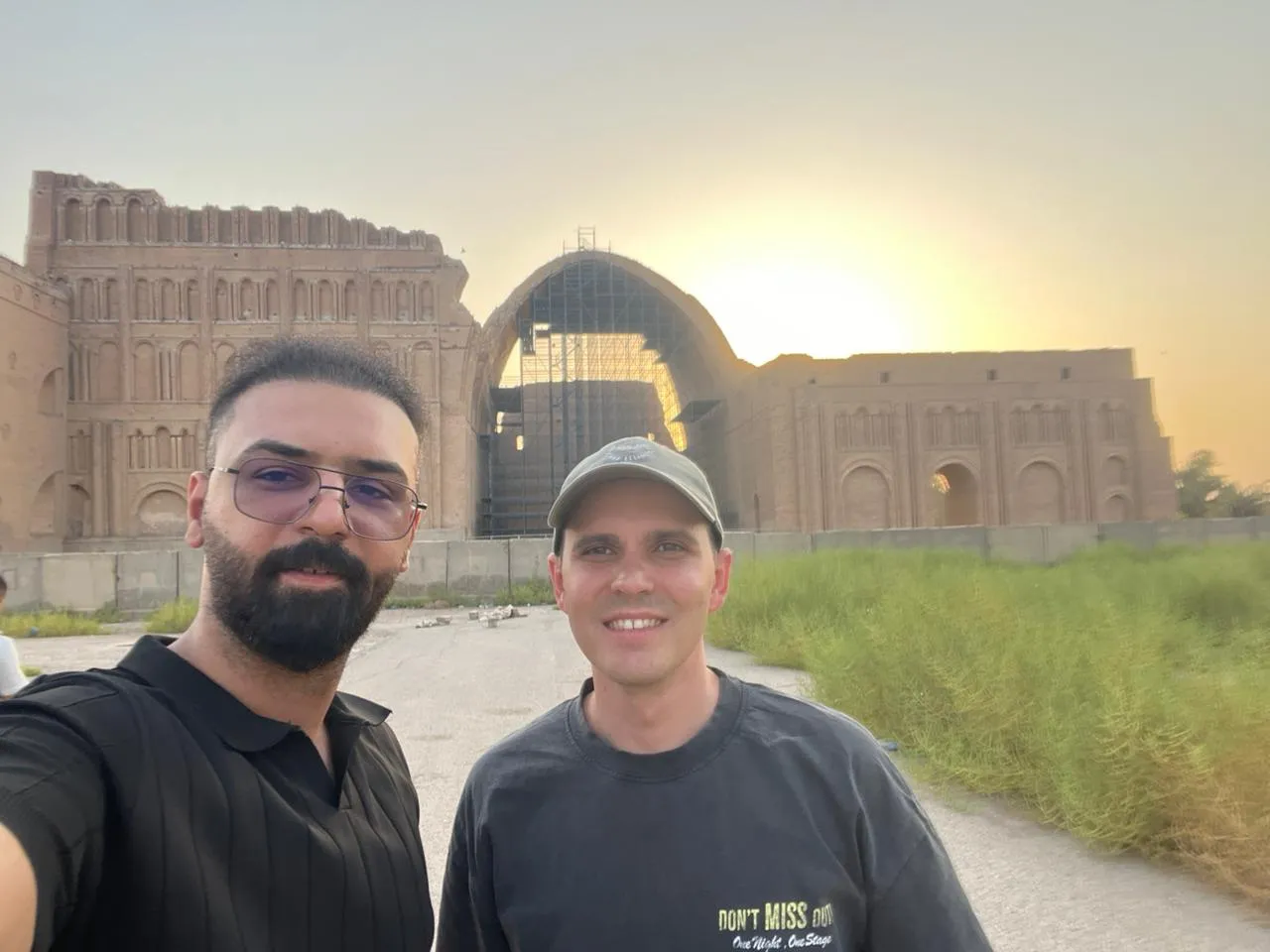 Two travellers at Ctesiphon with the brick vault of Taq Kasra rising behind them in the last of the light.