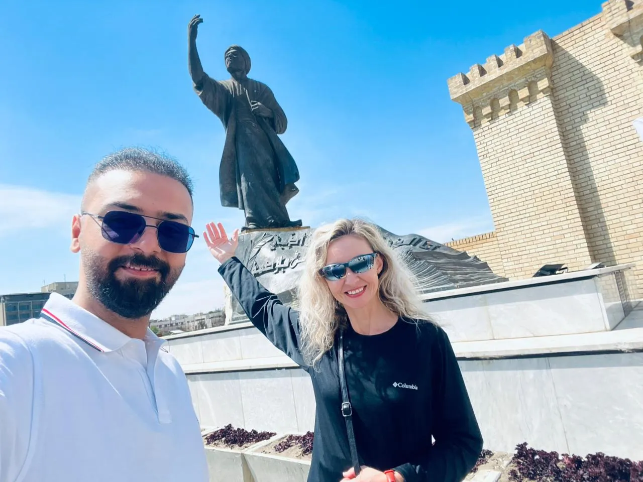 Two travellers below a statue at Erbil's airport welcome monument, its citadel-style wall running behind them.