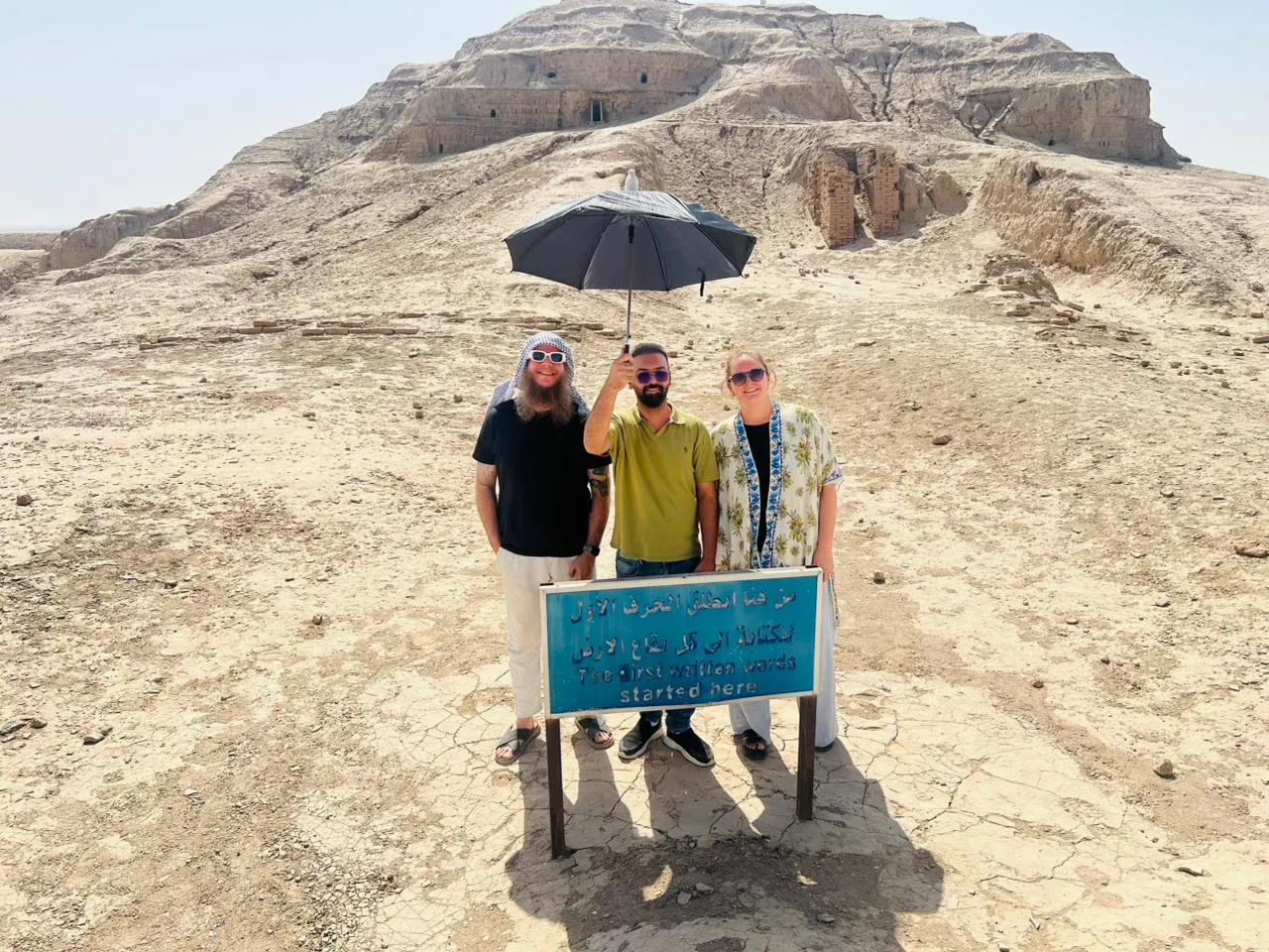 Three travellers sheltering under an umbrella on an excavation mound, beside a sign telling visitors that history started here.