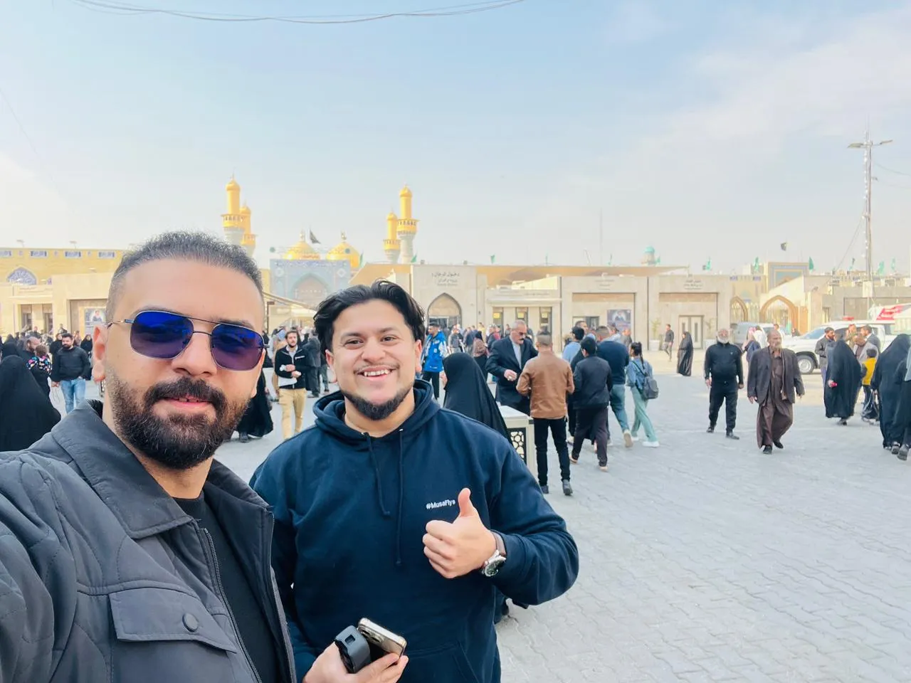 Two travellers on a busy shrine plaza, a golden dome and minarets rising through the crowd behind them.