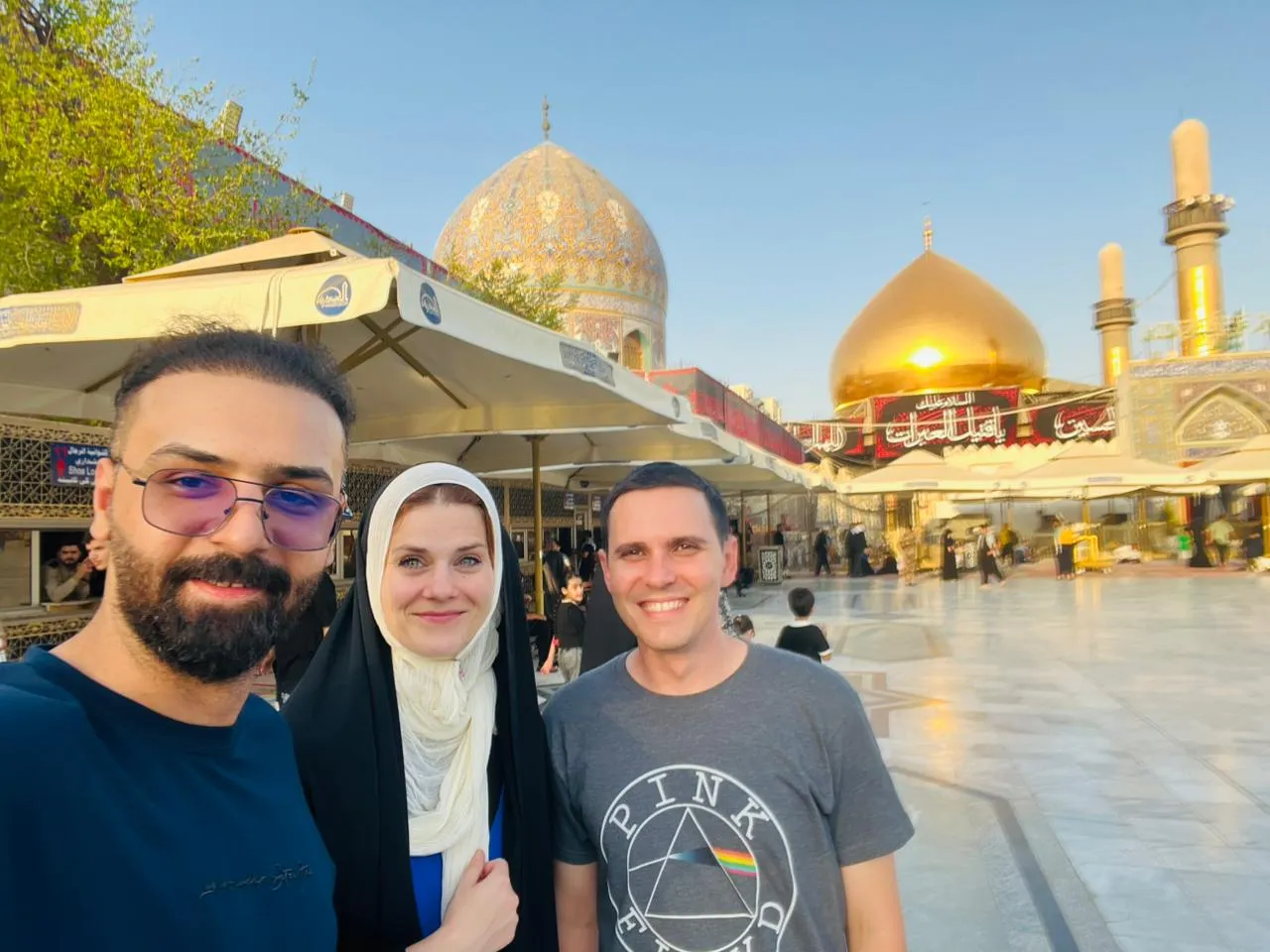 Three travellers at the edge of a shrine courtyard, the gold dome and tiled minaret catching the afternoon light.