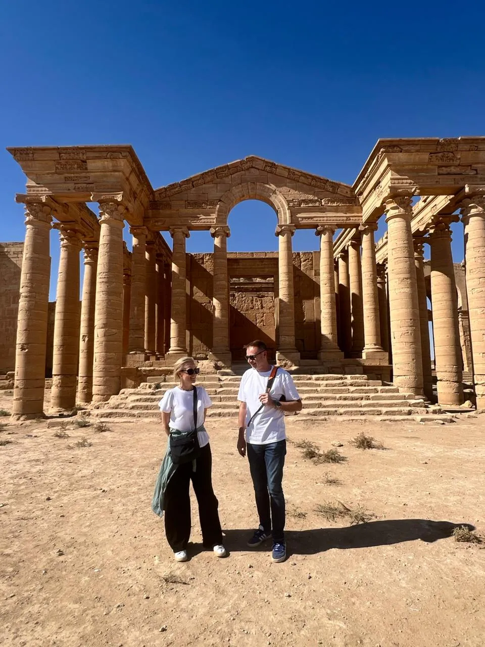 Visitors on the steps of the great columned temple facade at Hatra, honey-coloured limestone standing to full height against a clear desert sky.