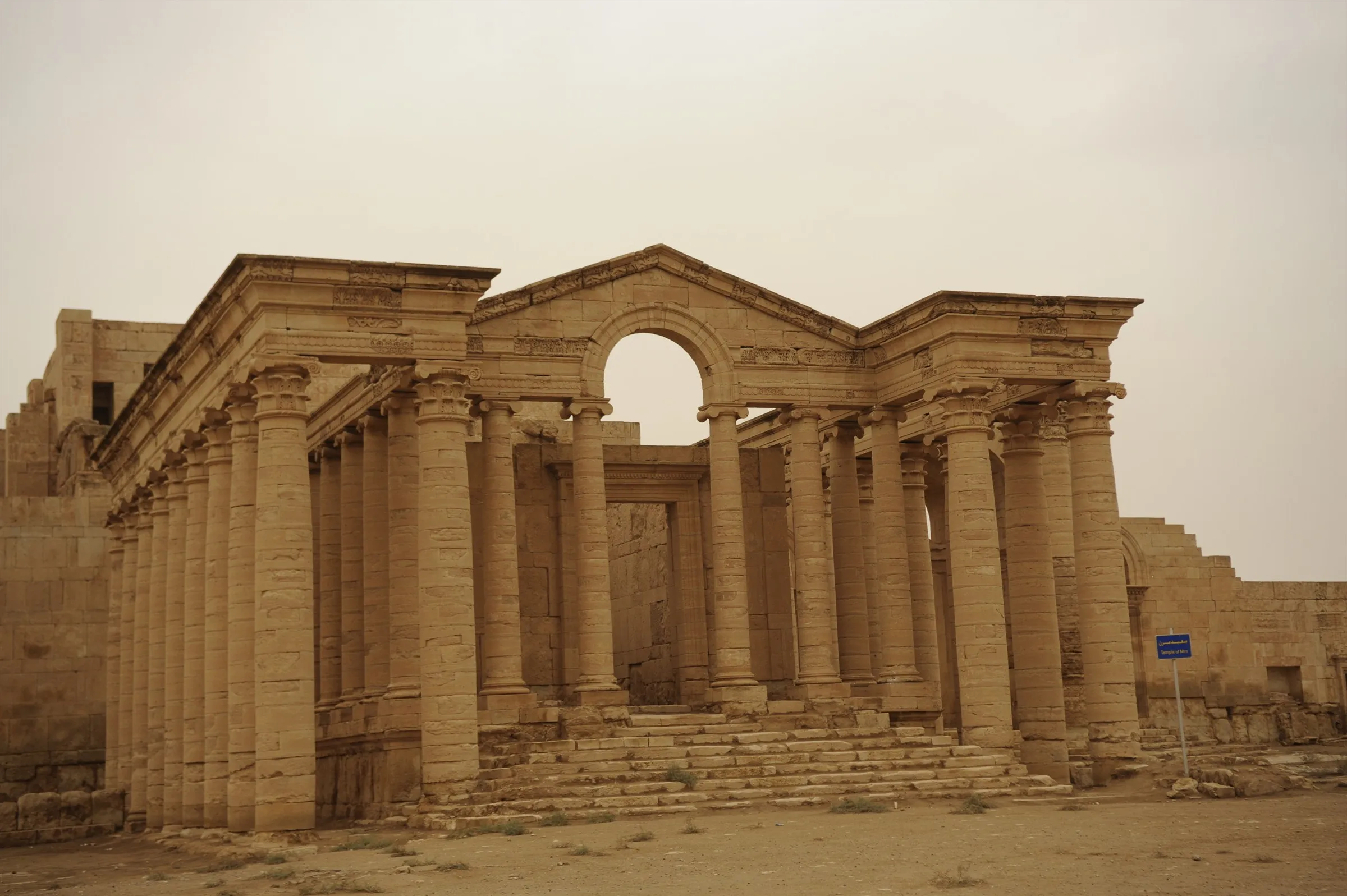 The temple facade at Hatra seen straight on, its pedimented Greco-Roman colonnade and arched centre standing intact against a pale desert sky.