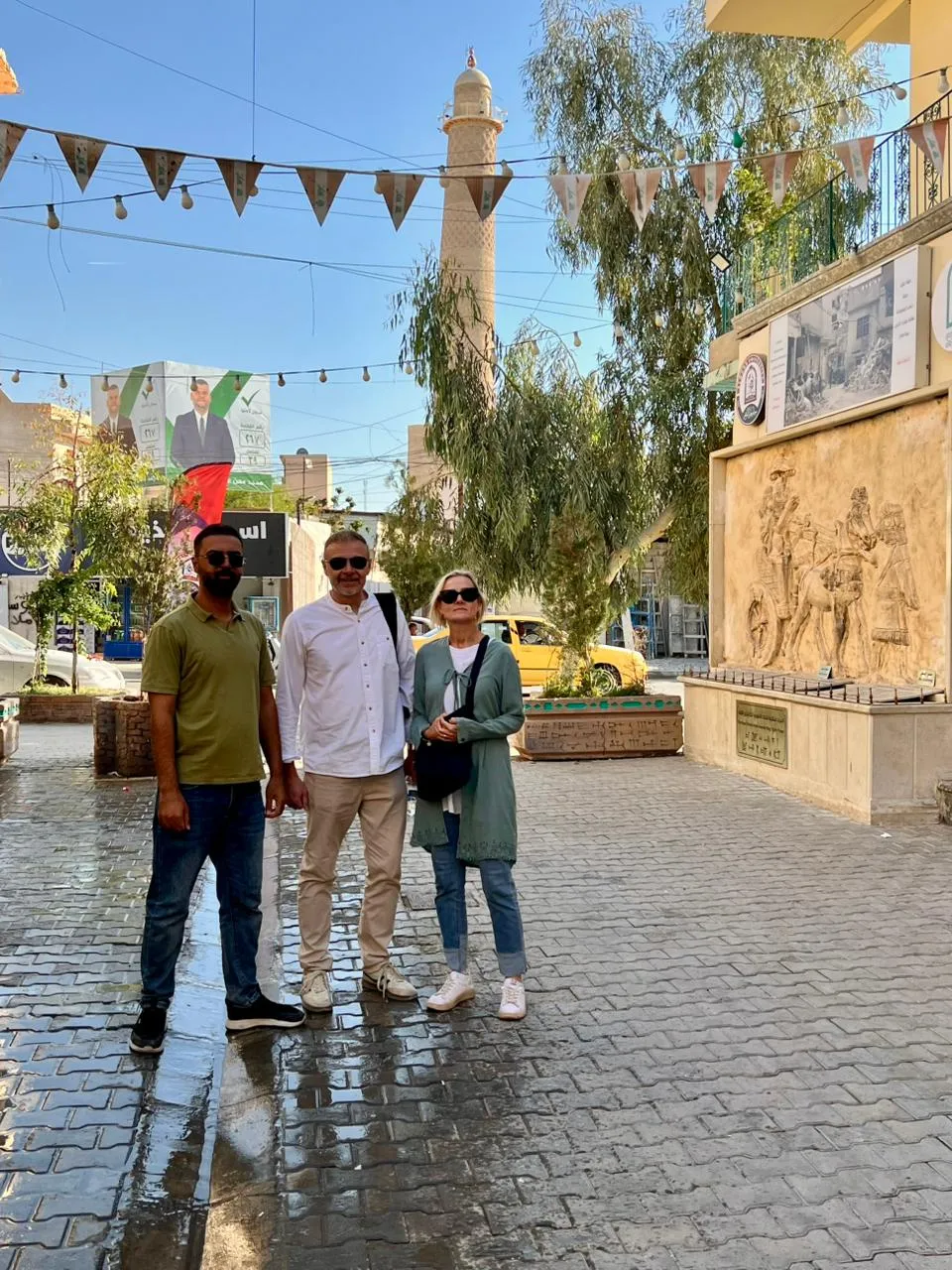 Three travellers on wet cobblestones in a heritage street, bunting strung overhead and a minaret beyond the trees.