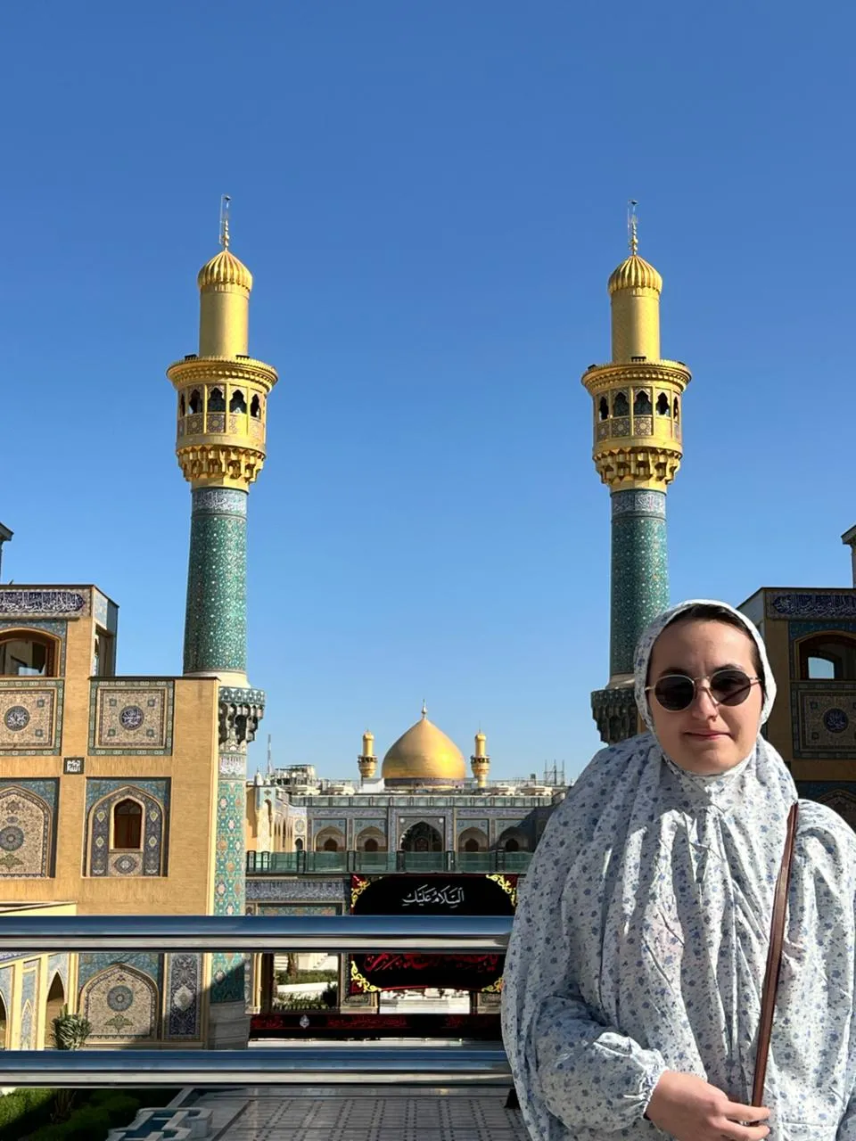 The gilded minarets and golden dome of the Kadhimiya shrine in Baghdad, a visitor standing before them in the forecourt.