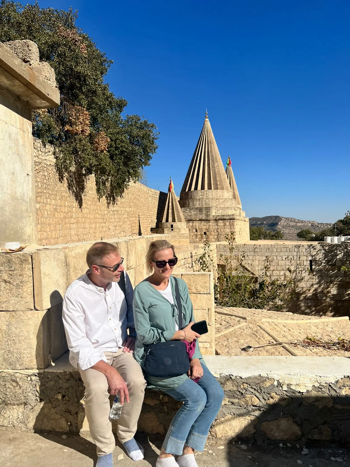 Two travellers sitting on a stone wall at Lalish, the fluted conical spires of the Yazidi temple rising beyond them.
