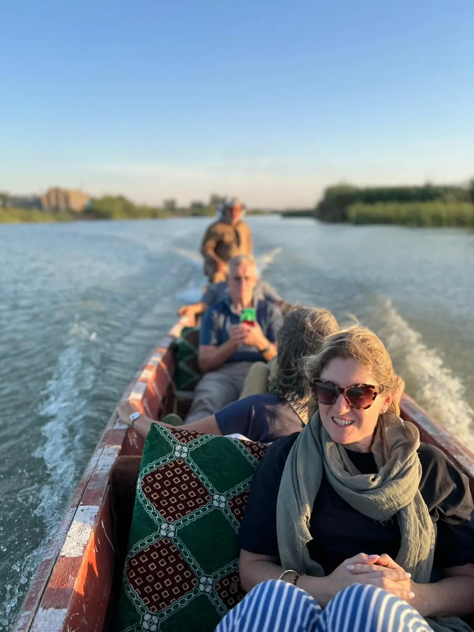 Travellers seated low in a long wooden boat running down a marsh channel at golden hour, reed banks slipping past on either side.