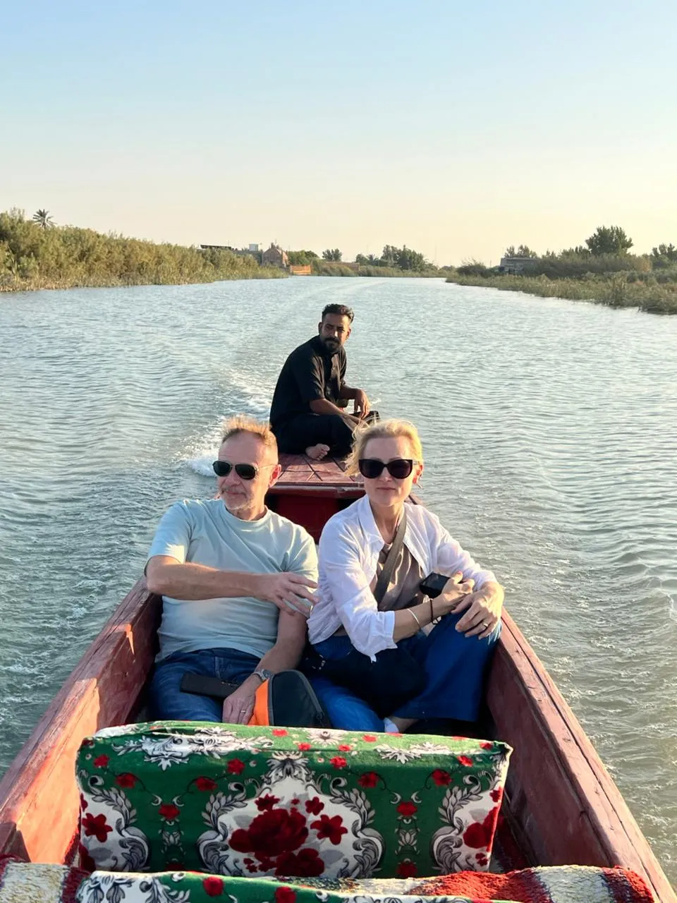 Visitors seated in a long wooden boat running down a southern waterway at golden hour, reed banks on either side and the boatman steering from the stern.