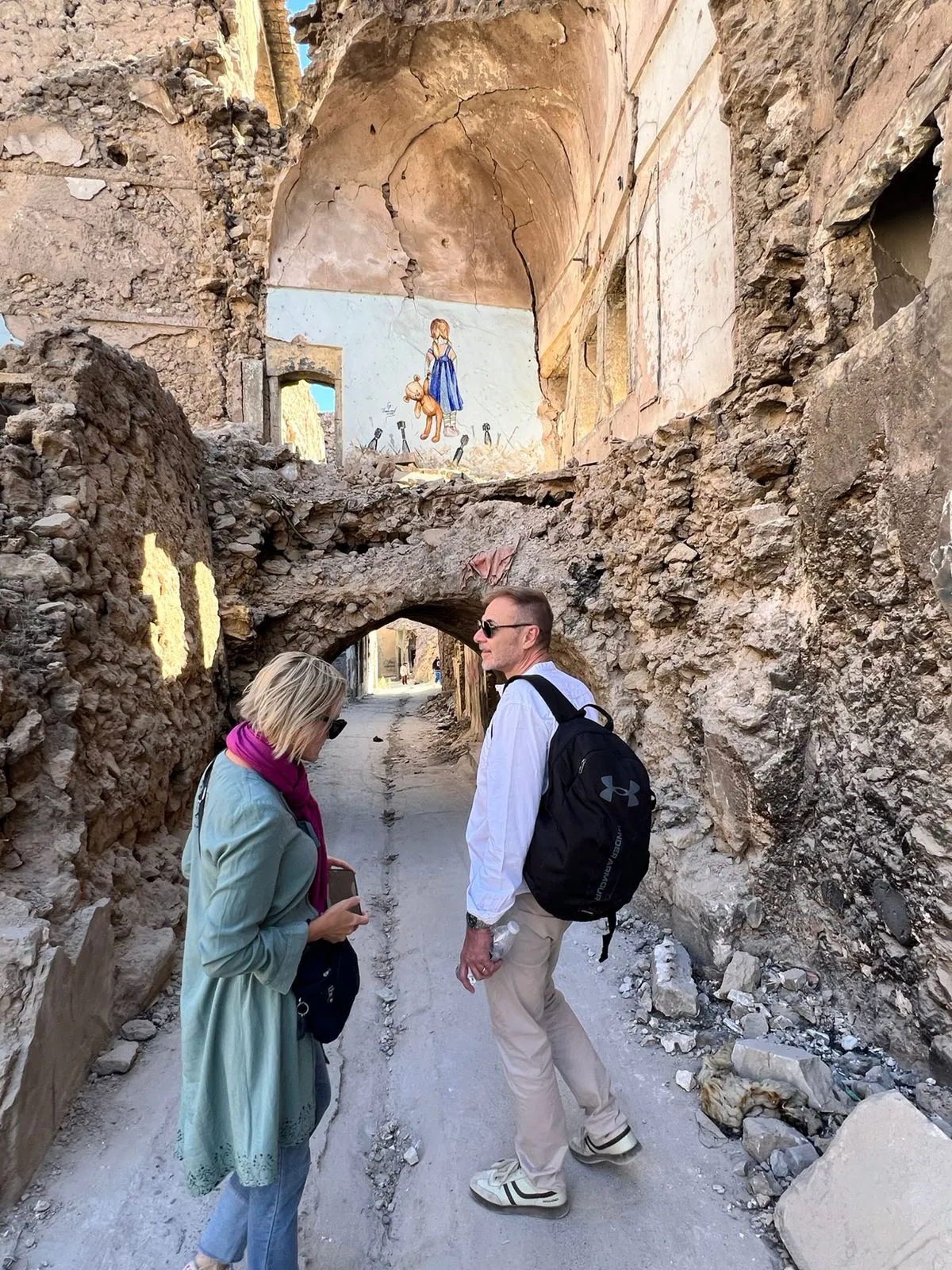 A vaulted alley in Mosul's old city, damaged stone walls on either side and a painted mural of a girl above the arch.