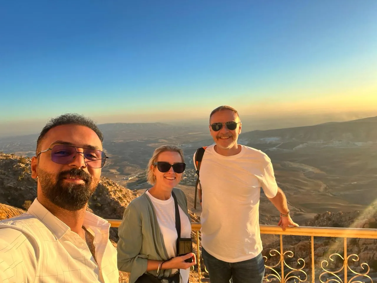 Three travellers at a railing on a northern mountain viewpoint, ridgelines fading into a hazy sunset.