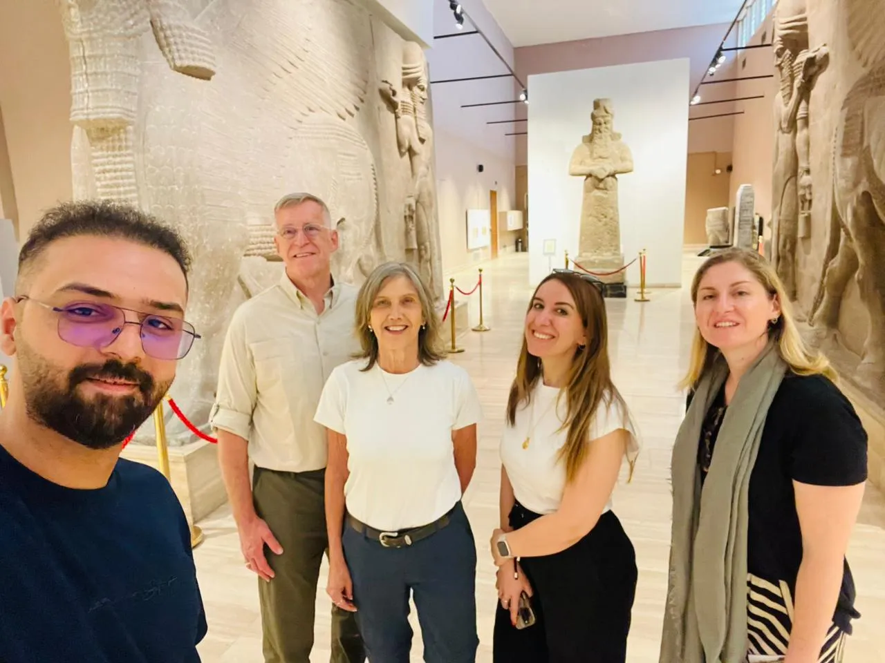 A group walking the Assyrian gallery of the Iraq Museum in Baghdad, carved reliefs and a standing figure lining the hall.