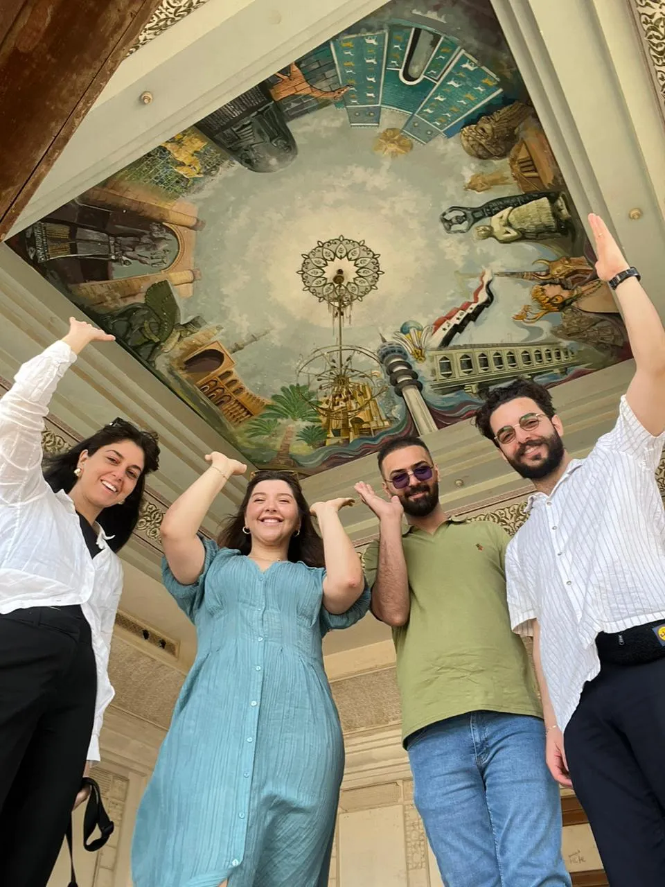 Four visitors under a painted ceiling, its panels crowded with scenes and a chandelier hanging at the centre.