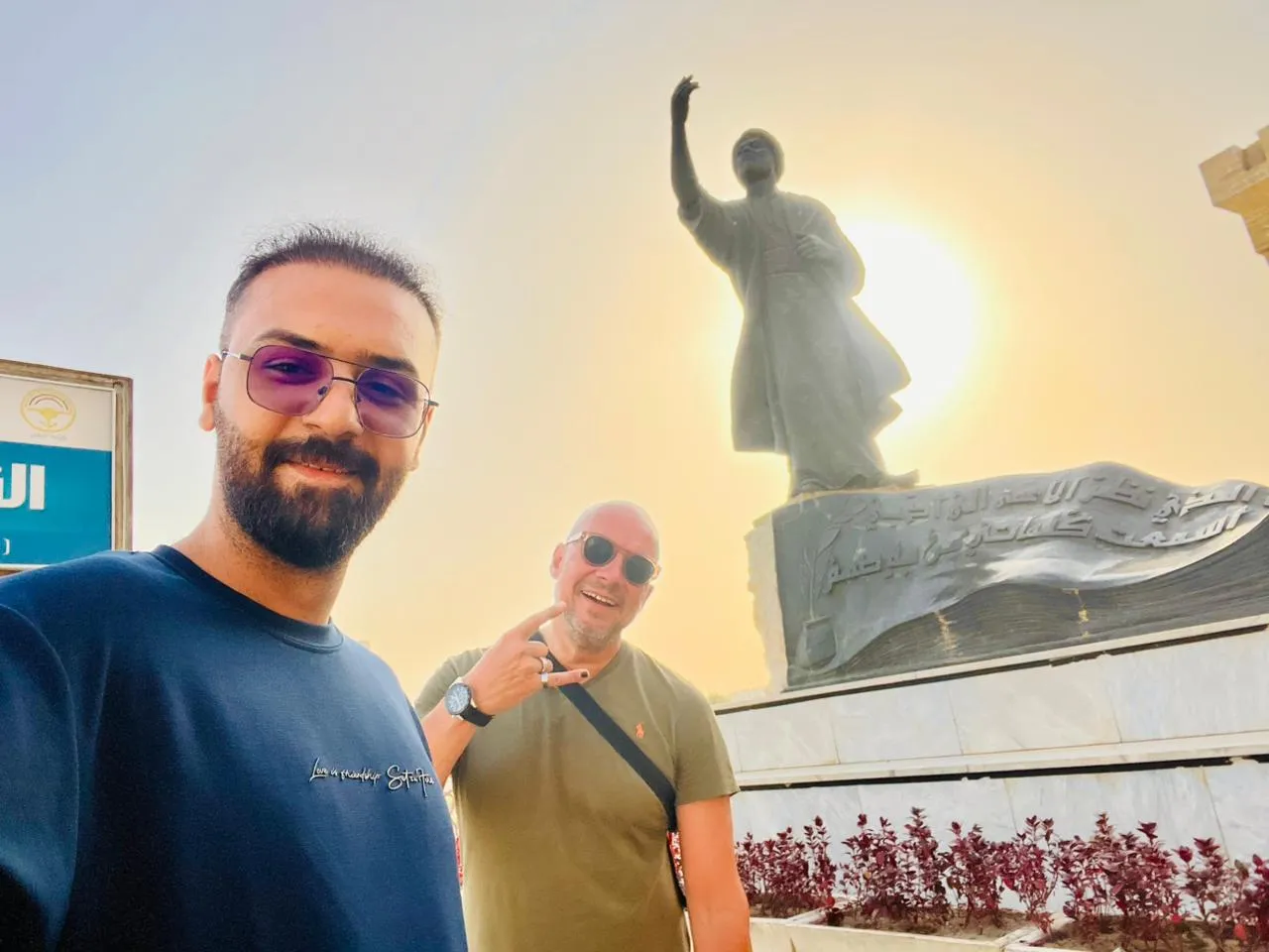Two men in front of a robed statue on a plinth, the low sun burning directly behind it.