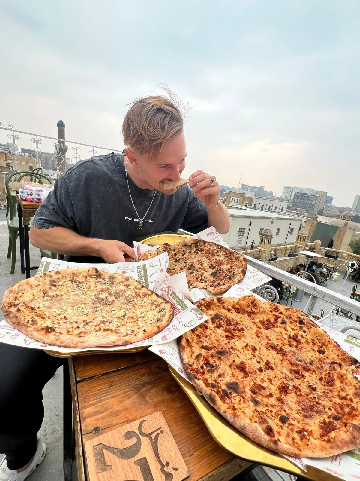 A traveller at a rooftop table with three flatbreads straight from the oven, the city roofline behind him.