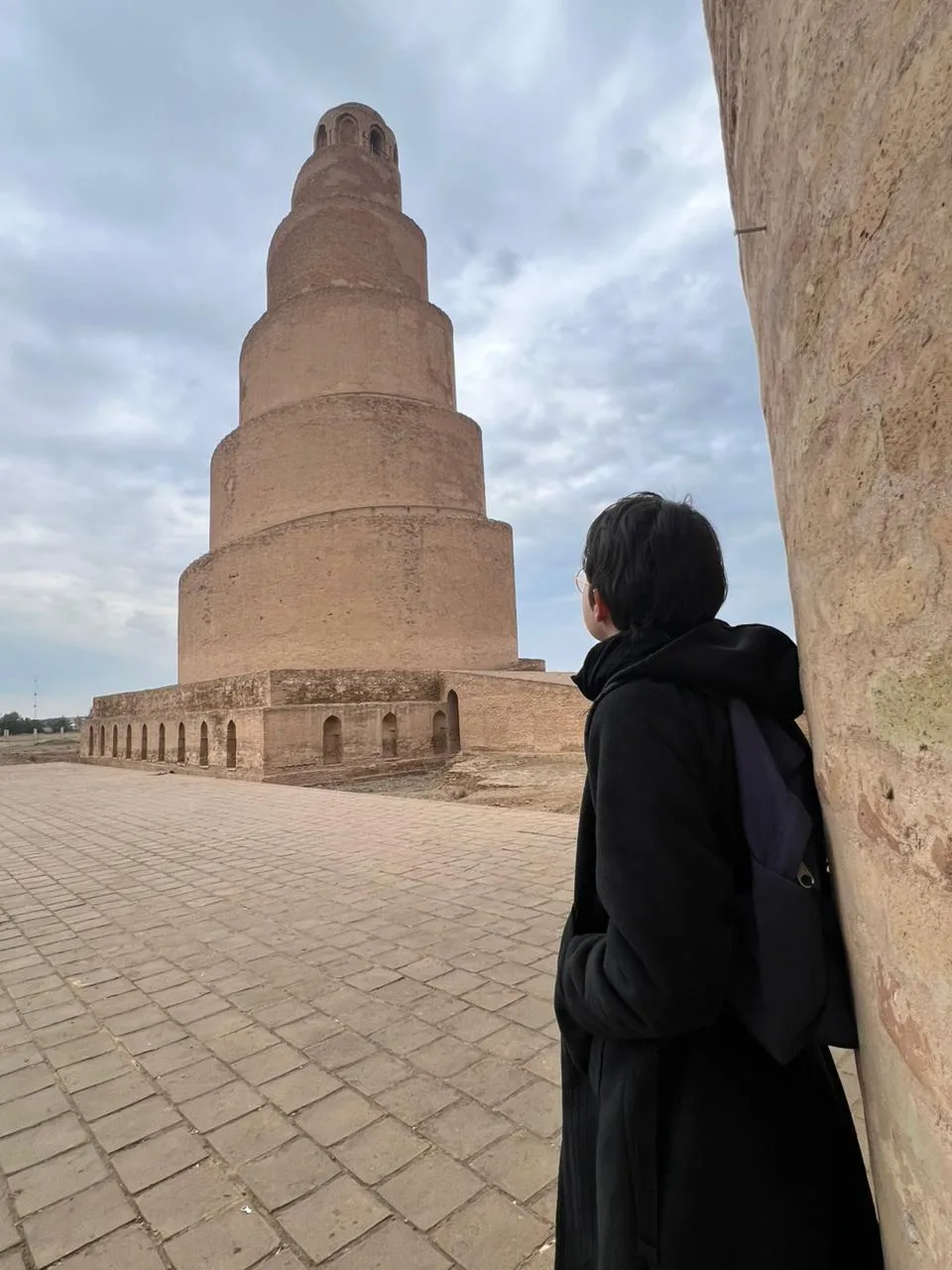 A visitor looking up at the spiral Malwiya minaret of Samarra from the shadow of the enclosure wall, the brick tiers of the tower rising beyond the arcade.