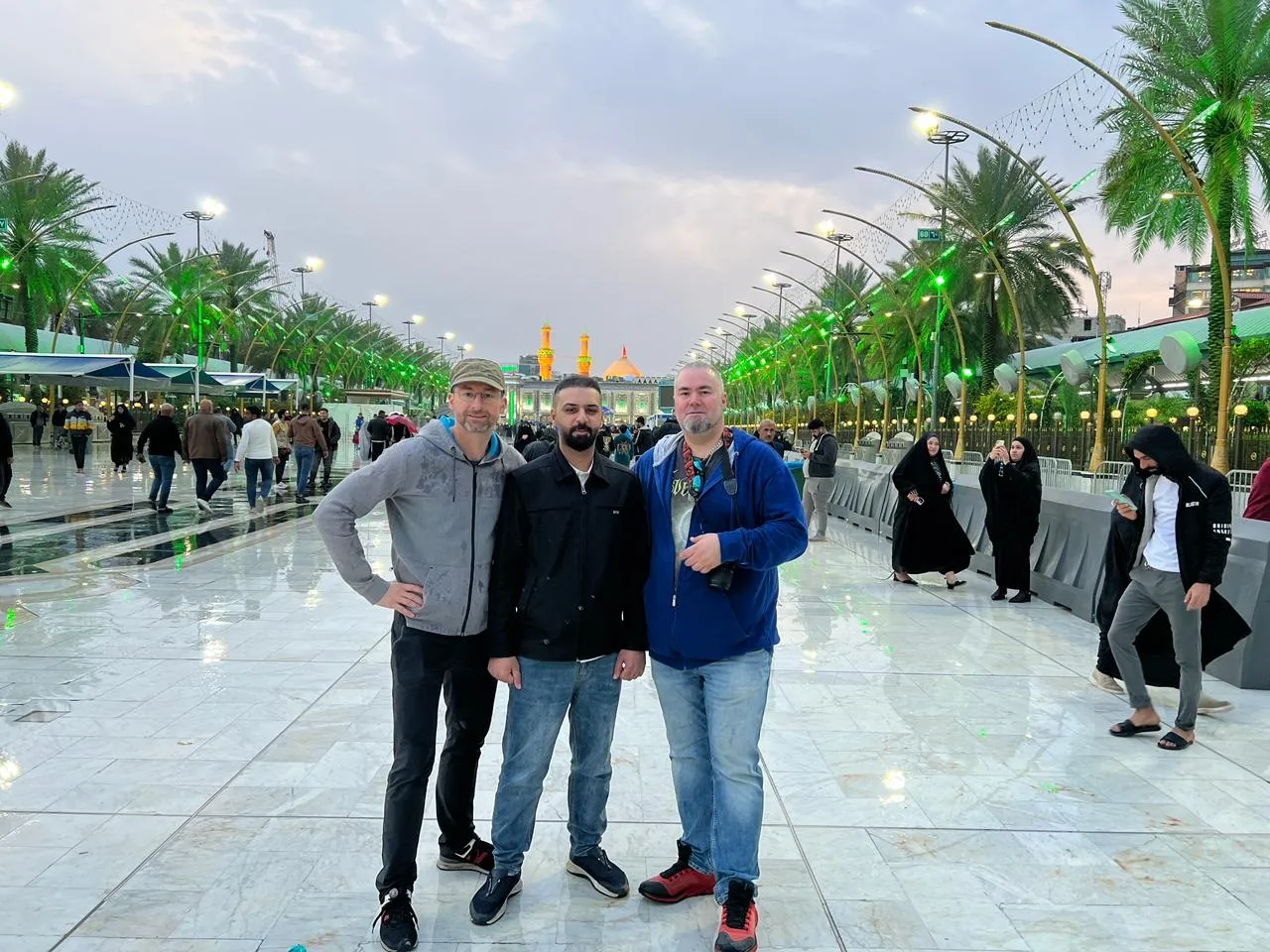 Three travellers on a wide shrine esplanade in the evening, banners and palms running away down its length.