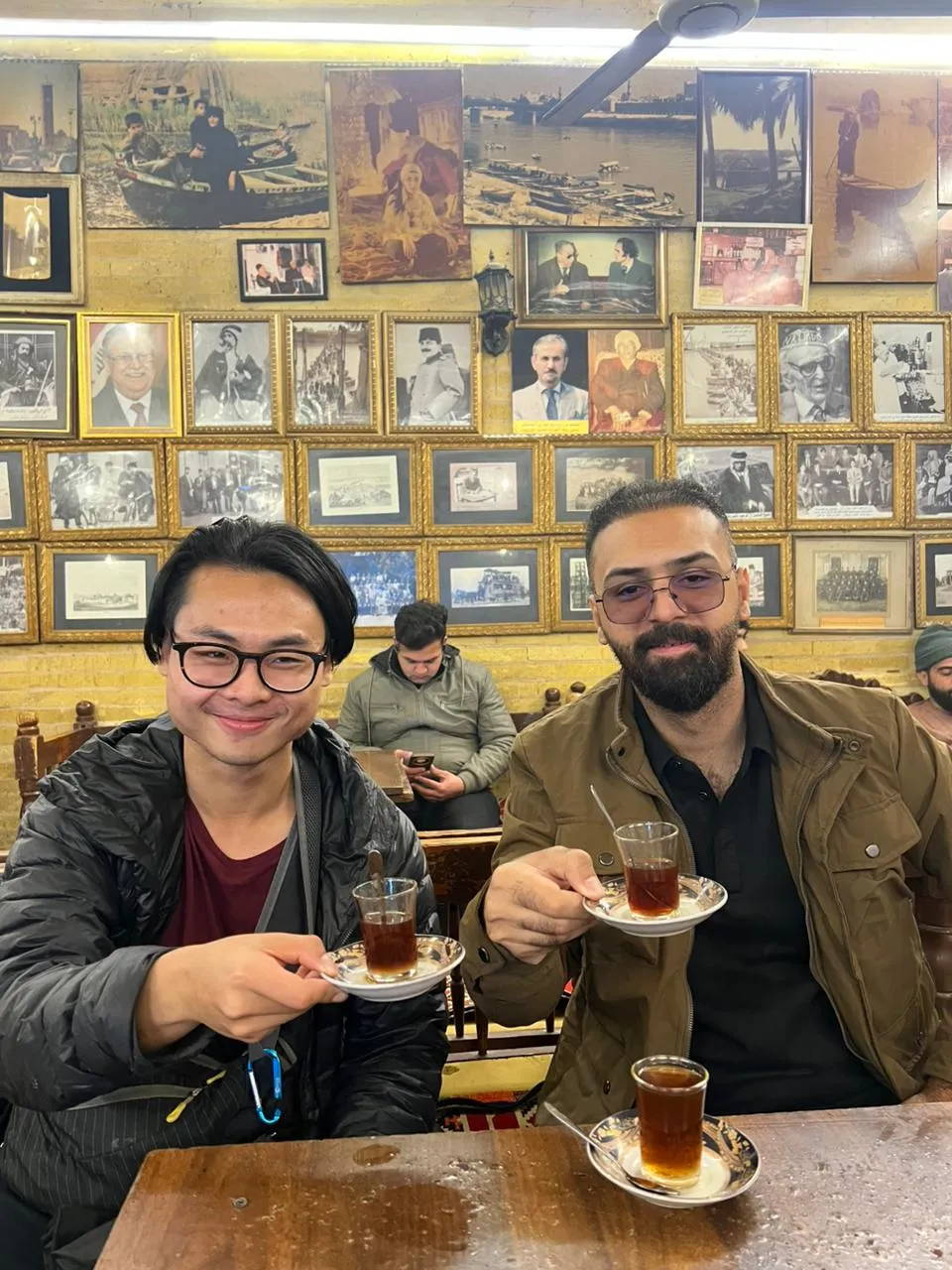 Two visitors raising istikan glasses of tea in a Baghdad tea house, the wall behind them covered to the ceiling in framed photographs.